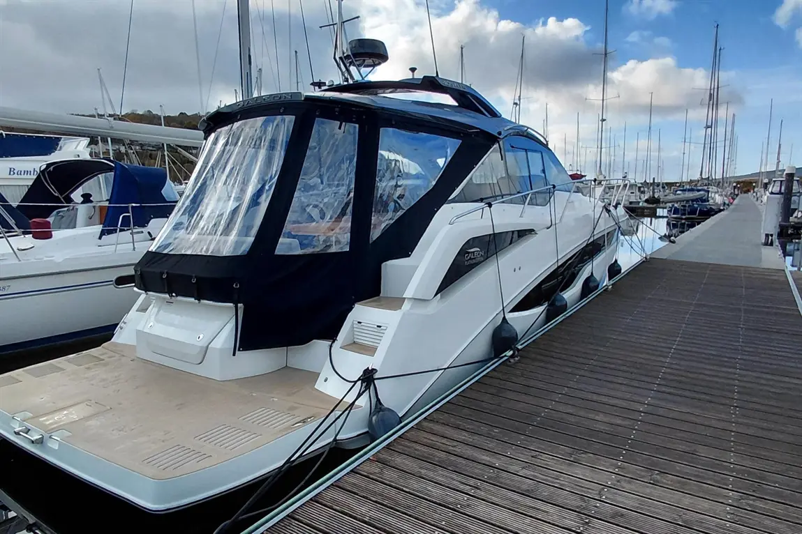 A sleek white yacht named "Galeon" docked at a marina, with wooden decking and other boats in the background under a cloudy sky.